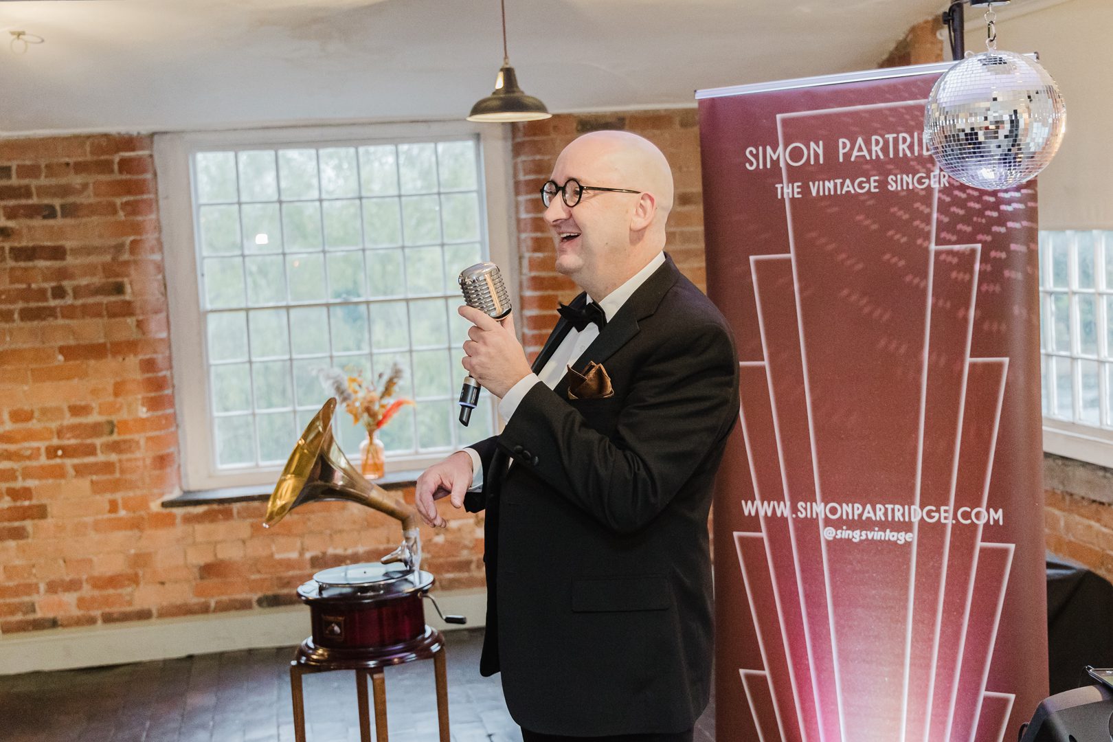 Singer in black tuxedo sings into vintage style microphone. Behind him is an art deco styled banner which reads Simon Partridge The Vintage Singer