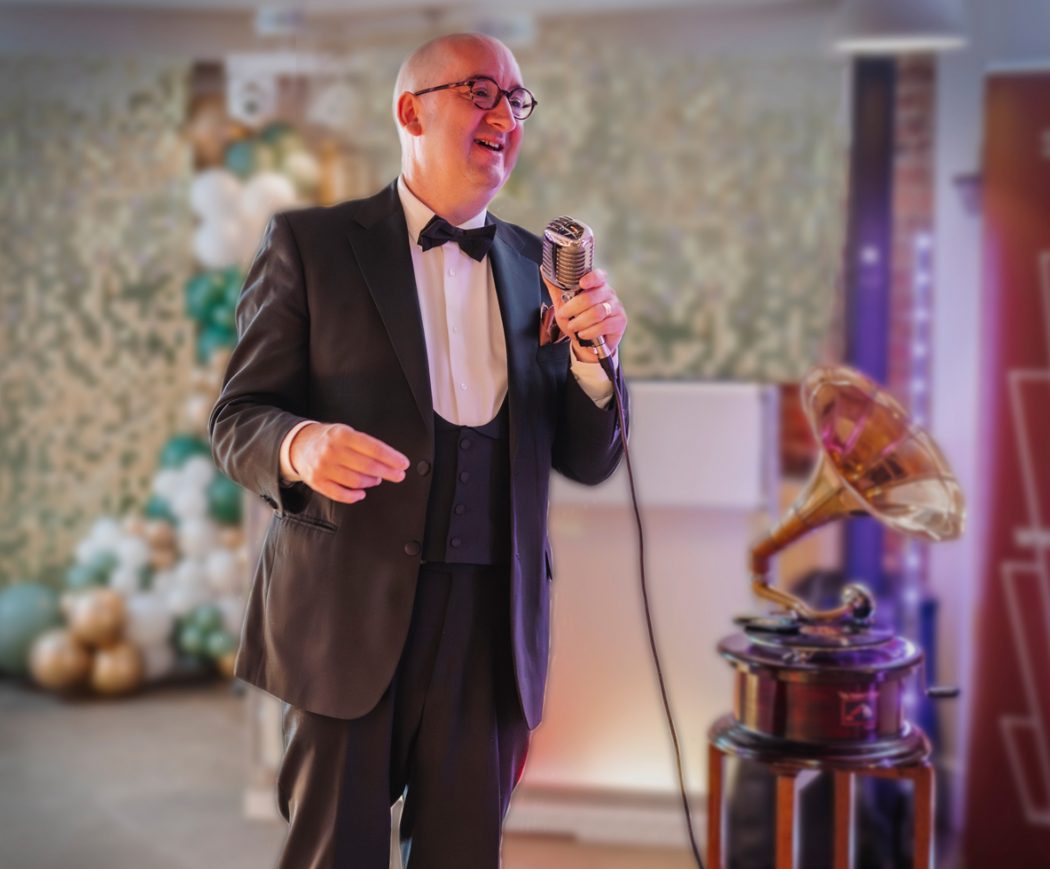 Singer in black tuxedo smiling. He hold a vintage style microphone and there is an old fashioned wind up gramophone next to him