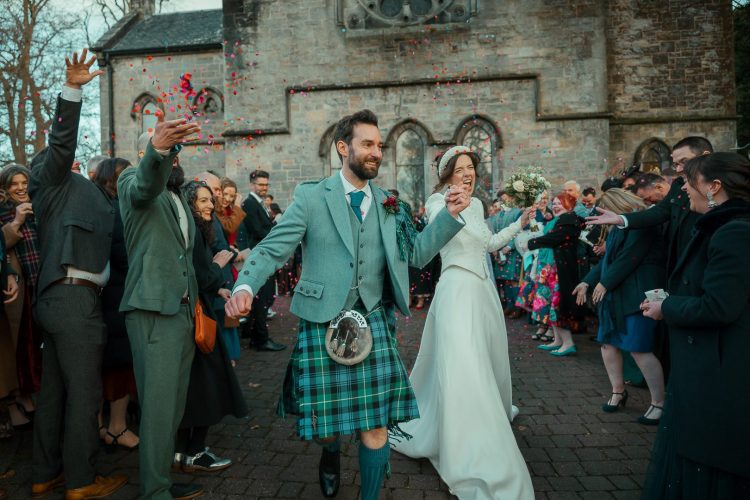 A Happy couple in green hues run under confetti with the church in the background