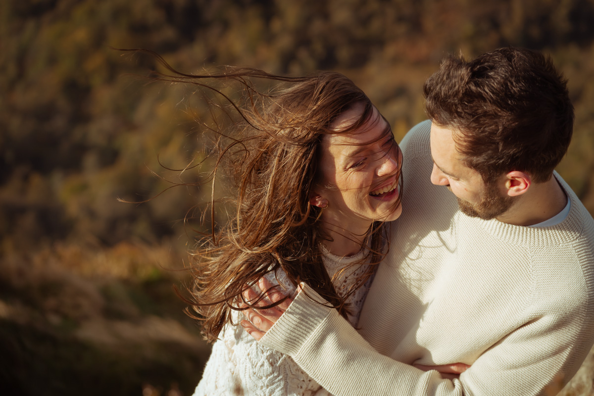 A windswept couple hold each other at the top of a mountain