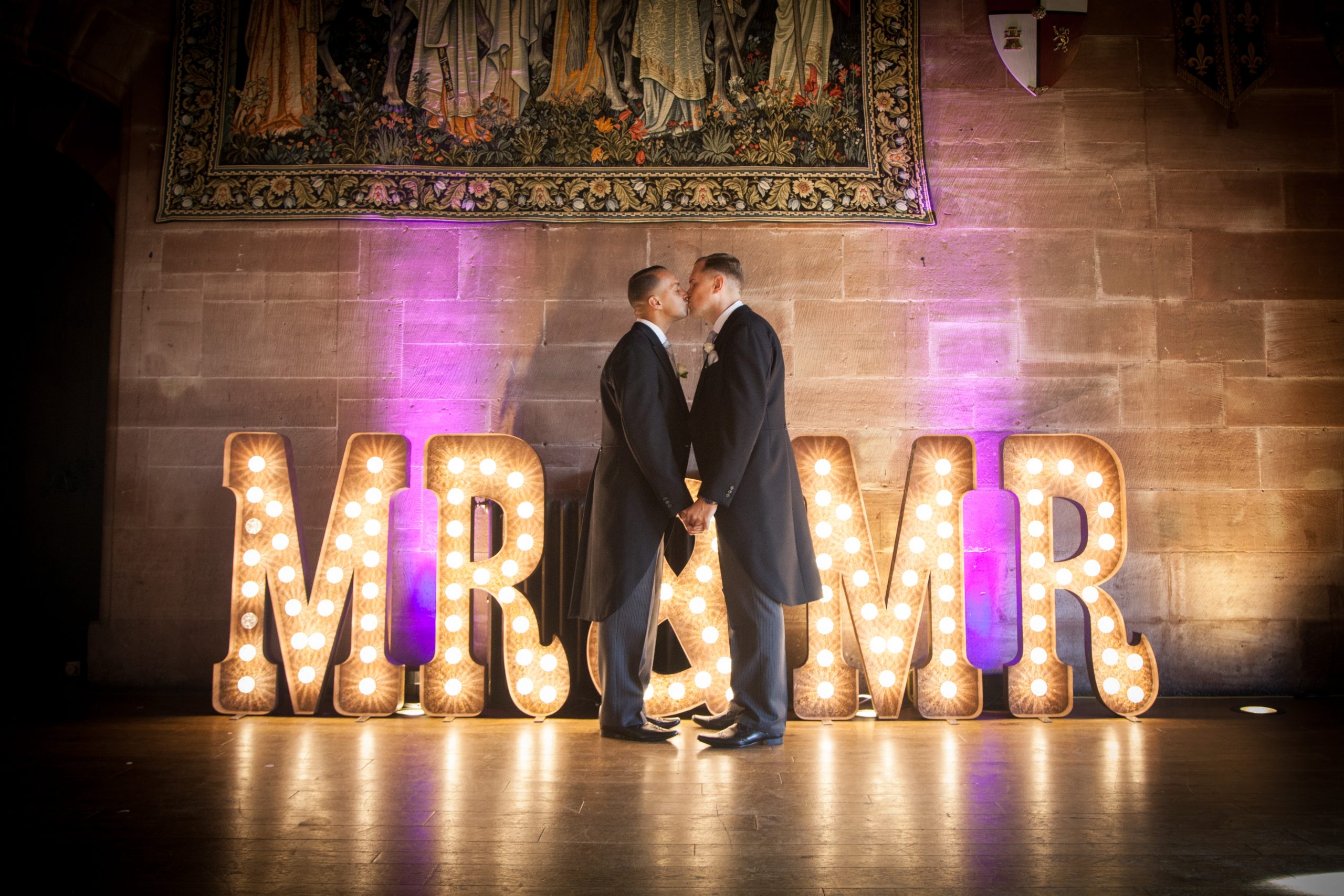 A same sex couple kissing while stood in front of a MR & MR illuminated sign