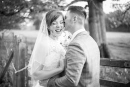 A black and white photograph of a couple, the groom is kissing the bride on the side of her head while she laughs.