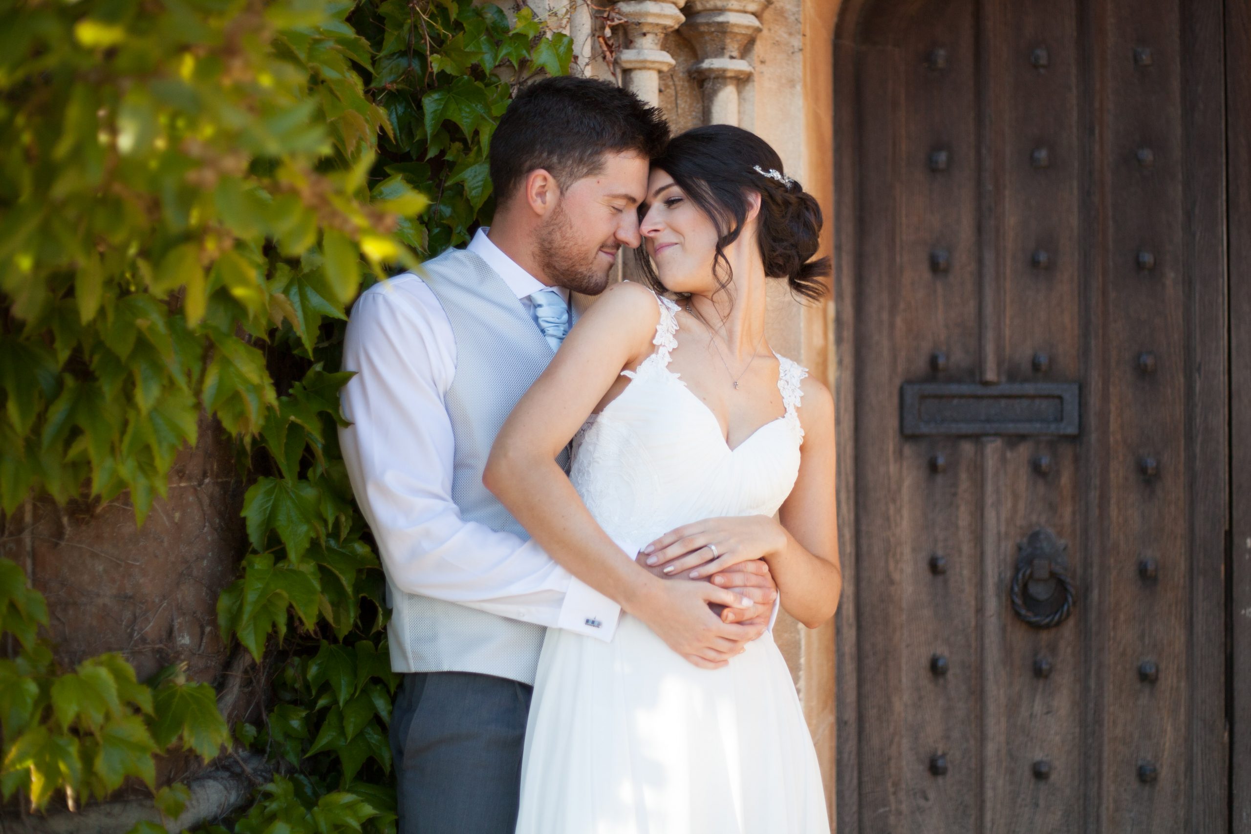 A romantic shot of a couple stood in front of an old doorway, with foliage to the side of them, they are leaning close into each other, romantically.