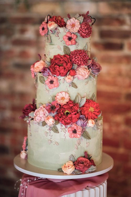 Three-tier sage green buttercream wedding cake decorated with red, coral and pink buttercream flowers