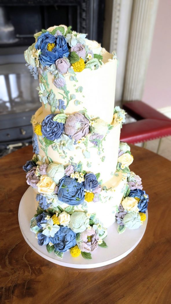 Three-tier ivory buttercream wedding cake covered with delicate blue, lilac and pastel buttercream flowers