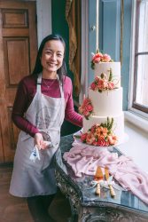The Flowering Baker beside a three-tier buttercream wedding cake decorated with coral and pink flowers